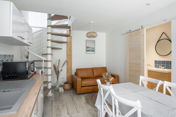 A modern kitchen with a built-in microwave, a dining area with a table and chairs, a brown leather sofa, and a staircase with wooden steps. - Le Coco de Cancale - Duplex proche mer (Cancale)