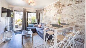 A modern living room with a sectional sofa, a dining table with chairs, a TV mounted on the wall, and a brick wall. - Galene - Appt avec jardin à 700 m de la Plage (Courseulles-sur-Mer)