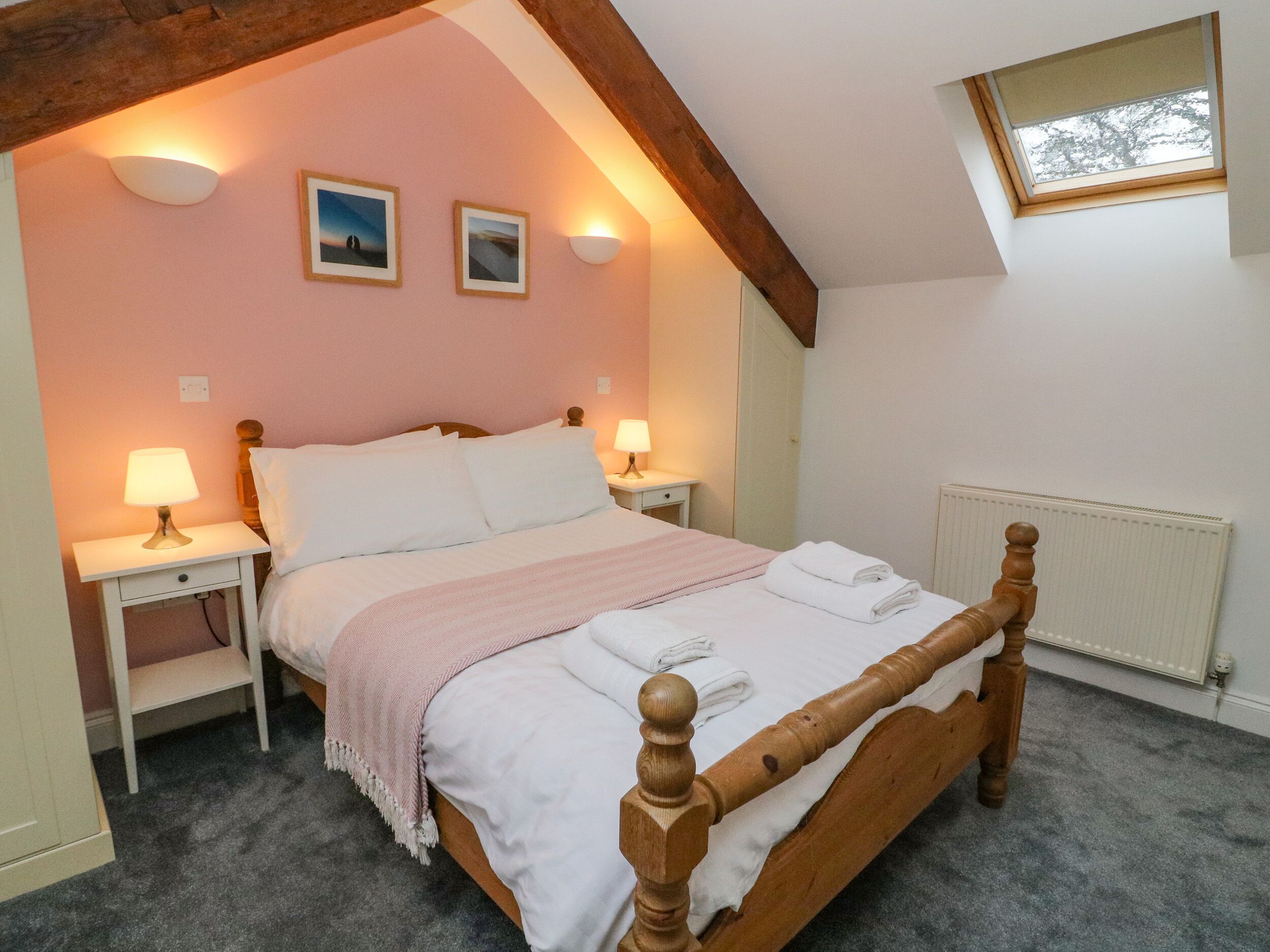 A bedroom with a wooden bed, two bedside tables with lamps, a pink wall, and a skylight.