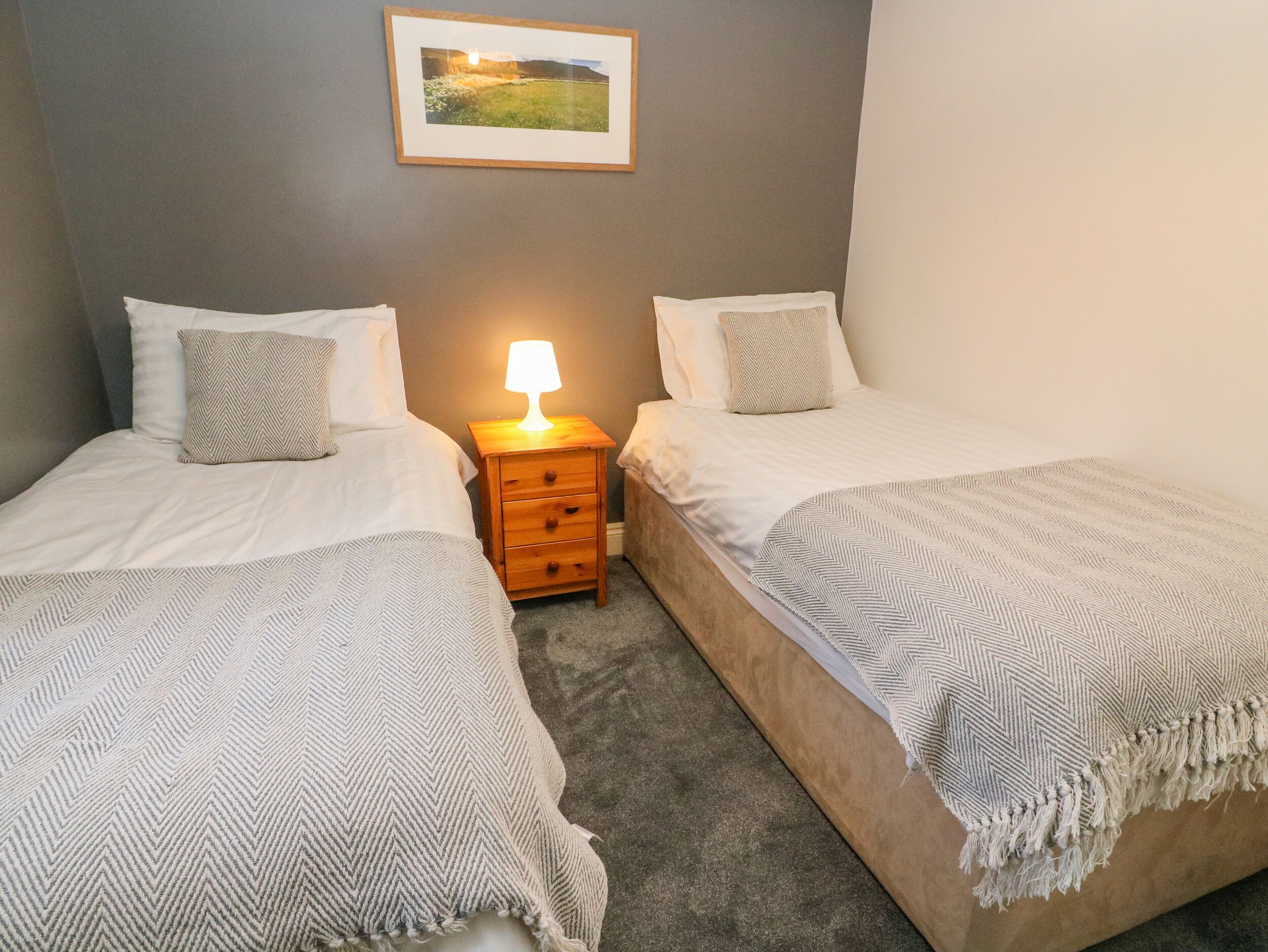 Quarto com duas camas, uma mesinha de cabeceira com abajur, um quadro pendurado na parede e piso coberto por carpete.