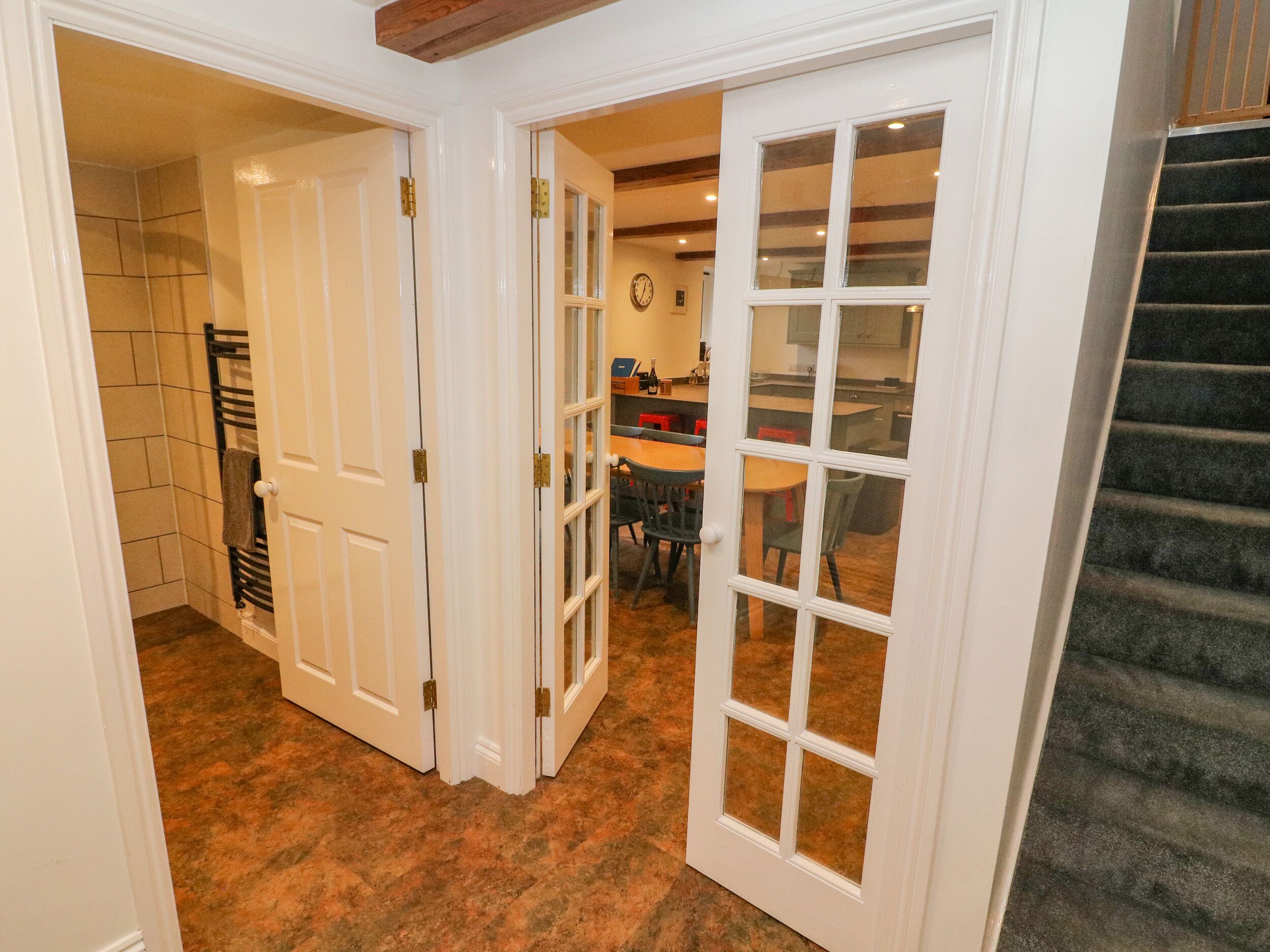A hallway with a staircase, a dining area with chairs and a table, and a kitchen area with cabinets and a clock.