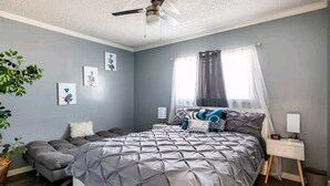 A bedroom with a bed, a bedside table, a nightstand, a ceiling fan, a window with curtains, and wall art.