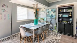 A dining area with a table and chairs, a clock, and a decorative shelf with plants.