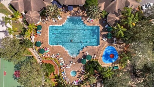 Ett flygbildsfoto av ett semesterområde med en central pool, solstolar, palmer och en basketplan.