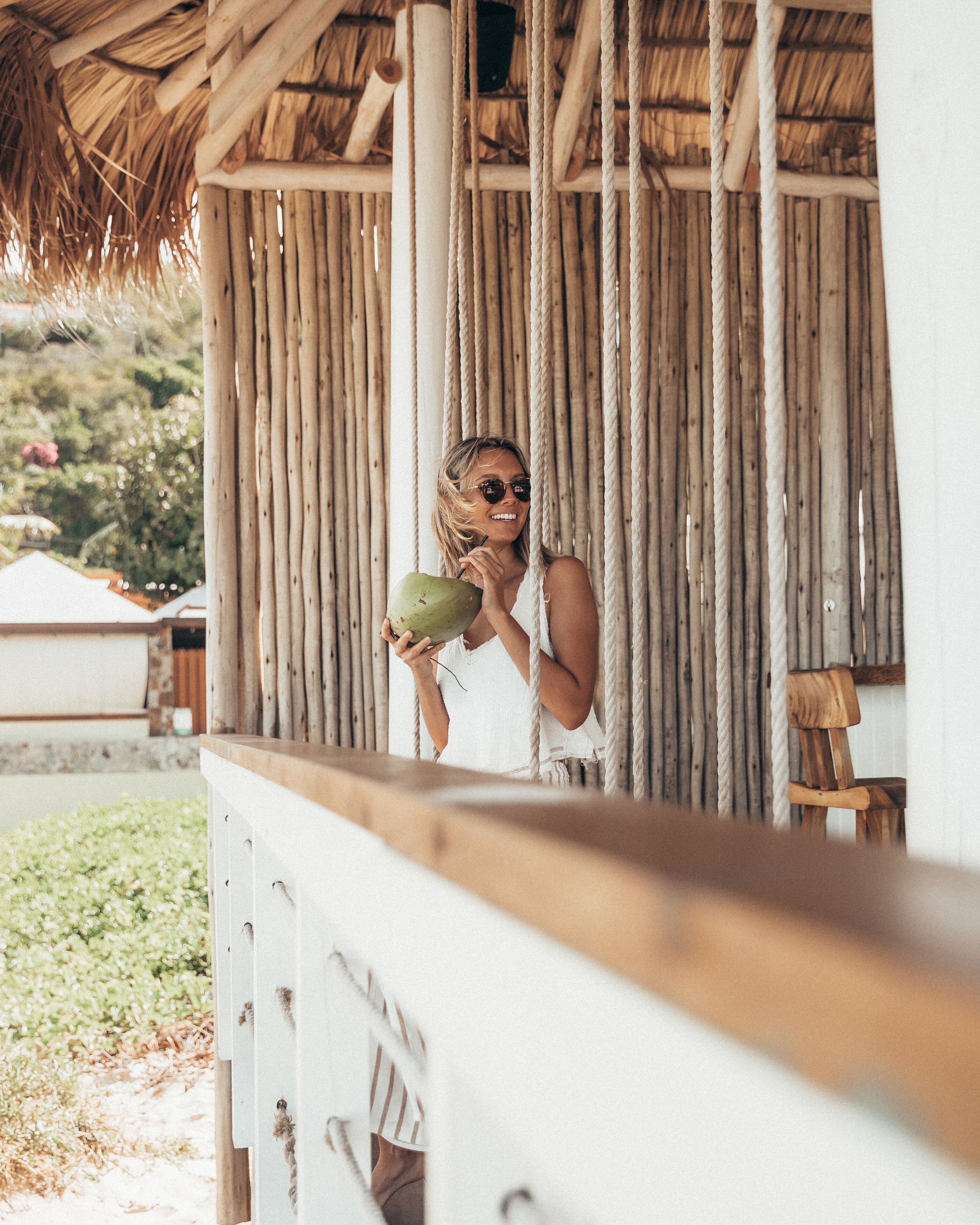 A person holding a coconut, standing under a thatched roof structure with vertical wooden beams.