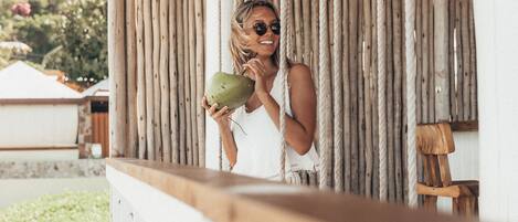 A person holding a coconut, standing under a thatched roof structure with vertical wooden beams.