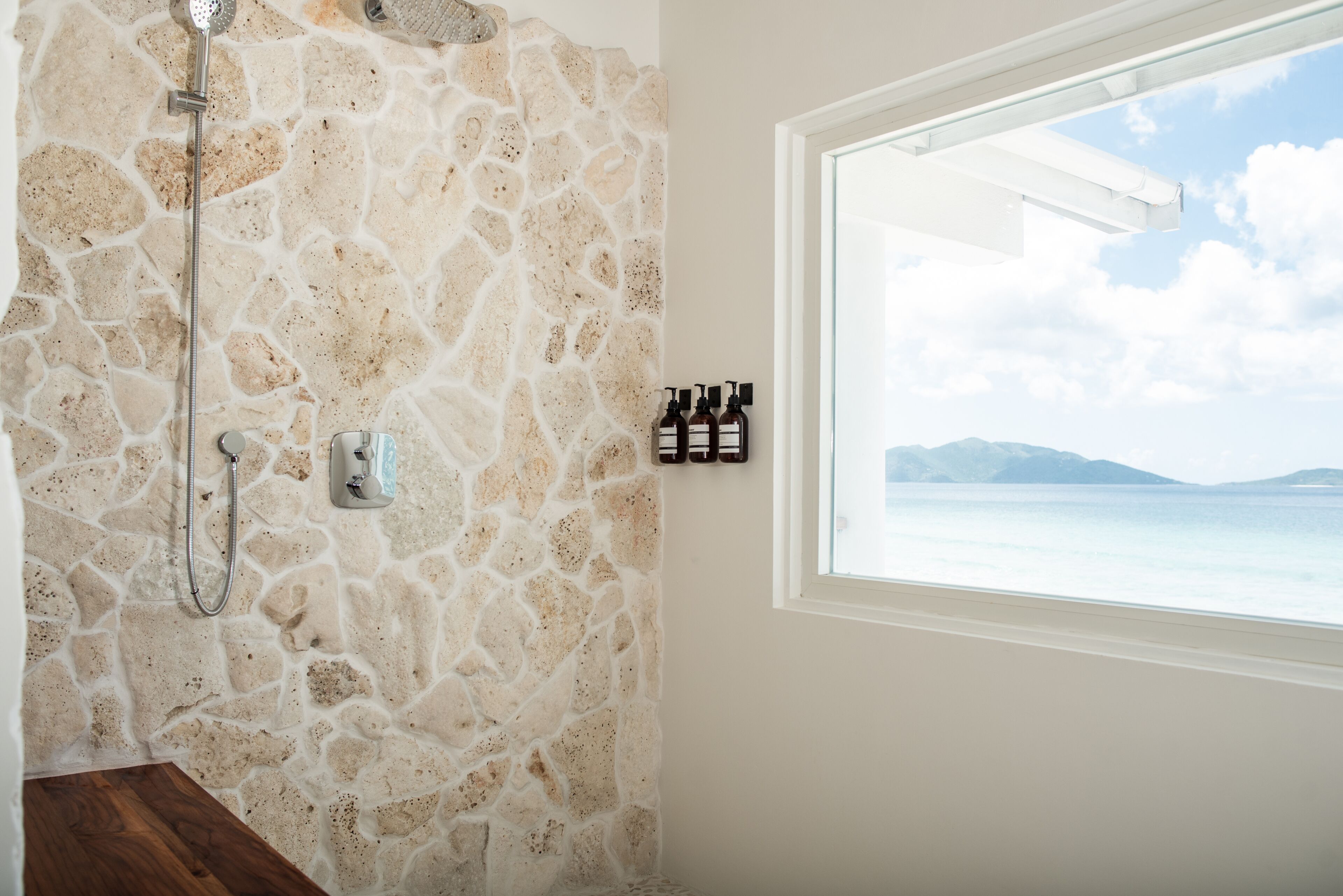 A bathroom with a stone wall, a shower, a window with a view of the sea, and toiletries on the wall.