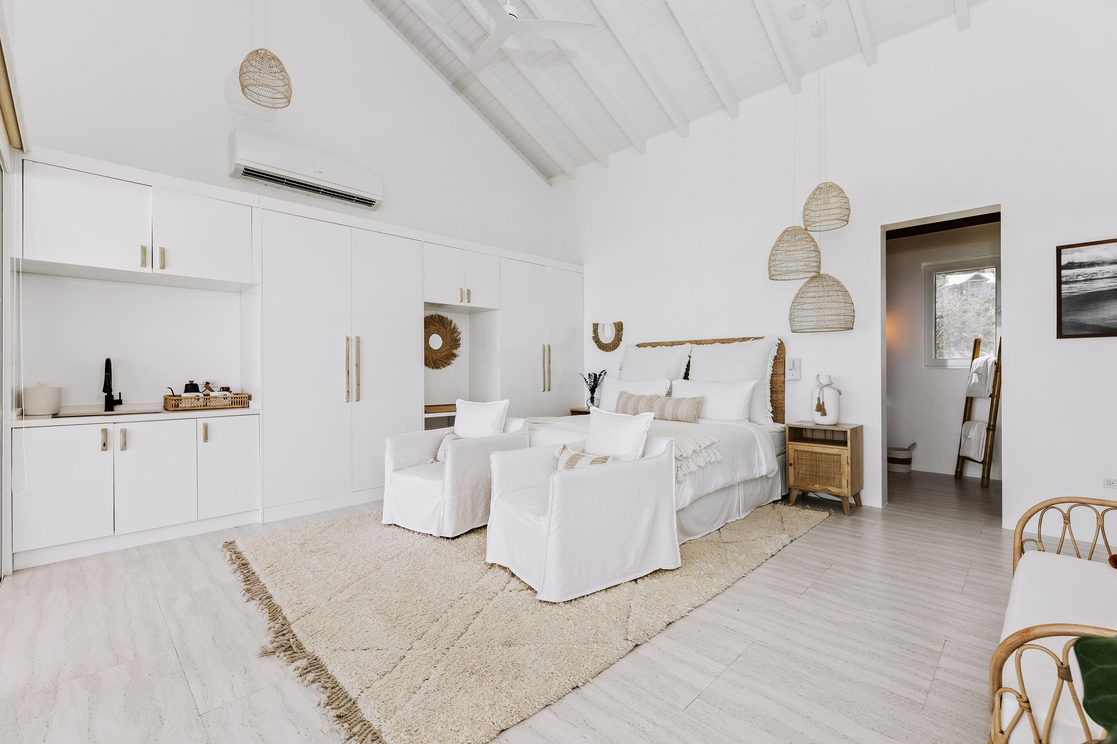 A spacious bedroom with a large bed, two white armchairs, a wooden nightstand, and a woven rug.