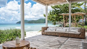 A covered outdoor seating area with a wicker sofa and a round table, overlooking a beach and ocean. - Long Bay Beach Resort (West End)