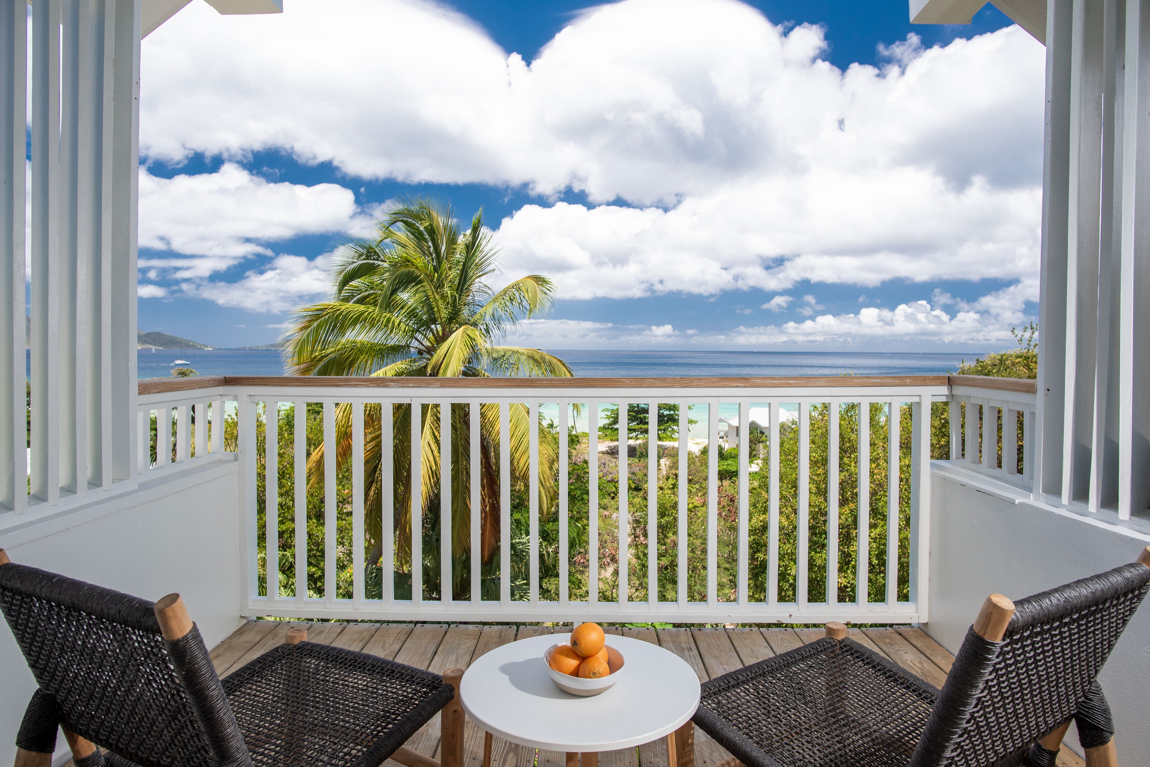 A wooden deck with wicker chairs and a table with fruit, overlooking a beach with palm trees and the ocean.