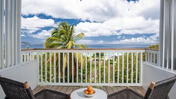 A wooden deck with wicker chairs and a table with fruit, overlooking a beach with palm trees and the ocean.