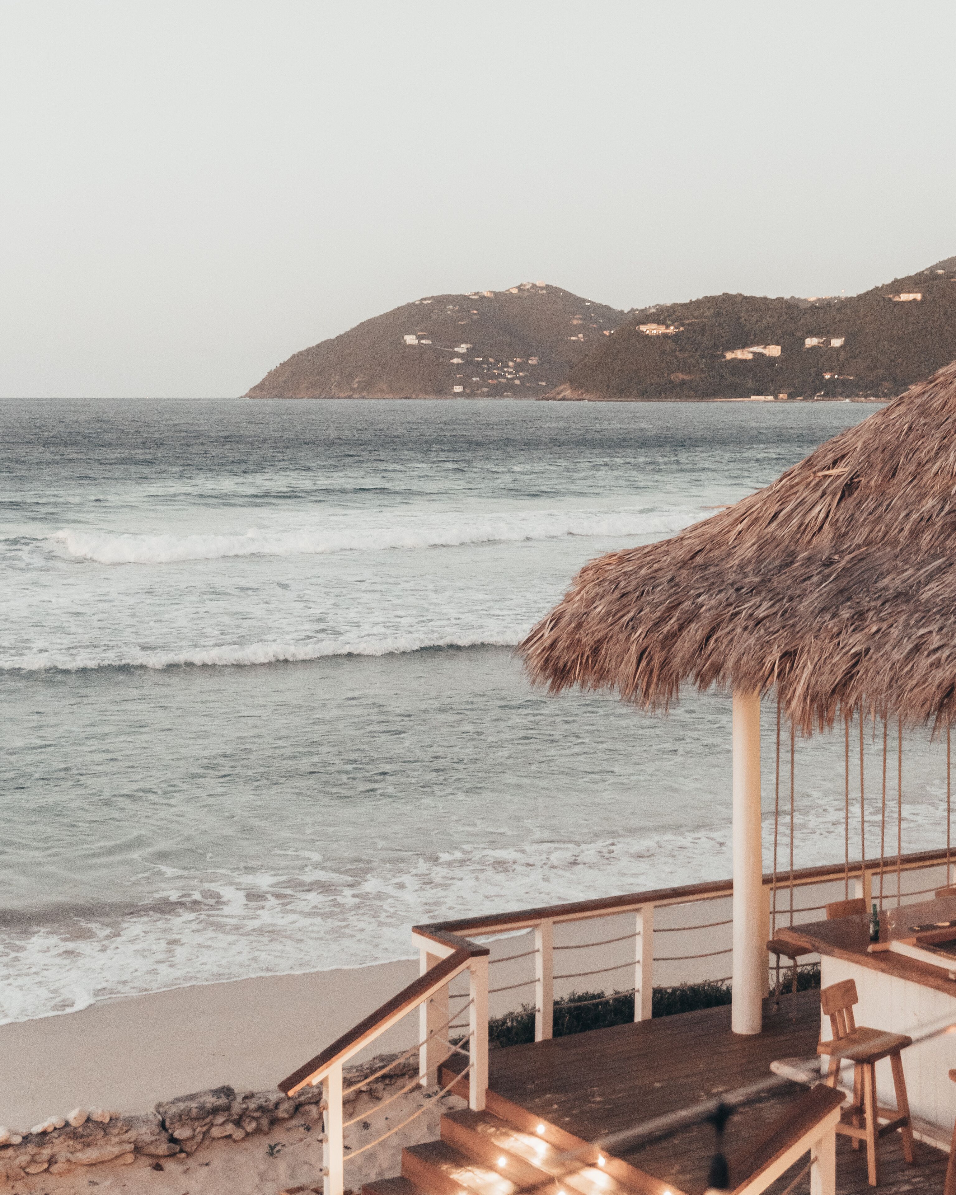 Une terrasse au bord de la plage, dotée d’un toit de chaume, d’une plateforme en bois et offrant une vue sur l’océan et les collines lointaines.