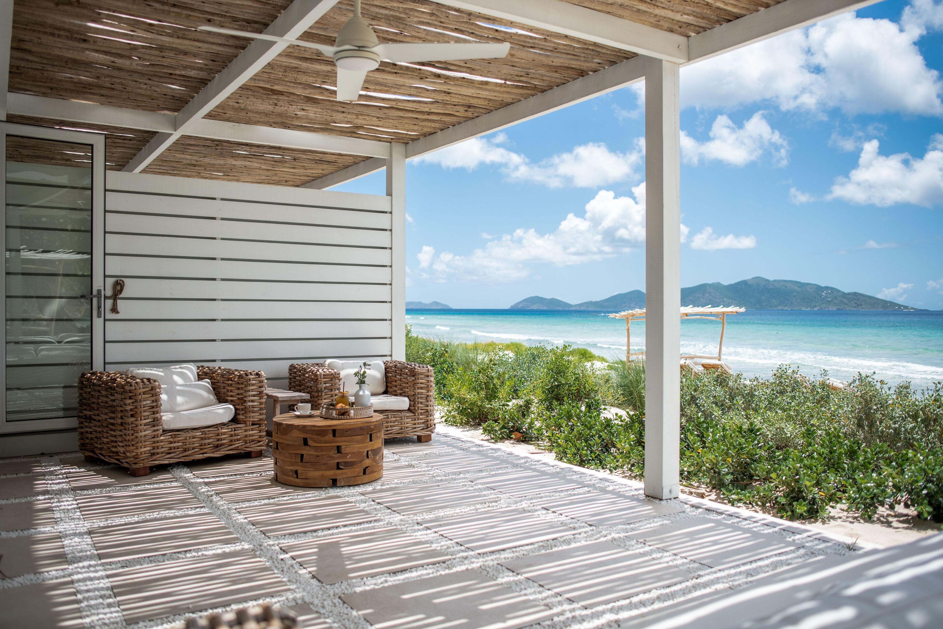 A covered outdoor seating area with wicker furniture and a wooden coffee table, overlooking a beach with clear blue waters and distant mountains.