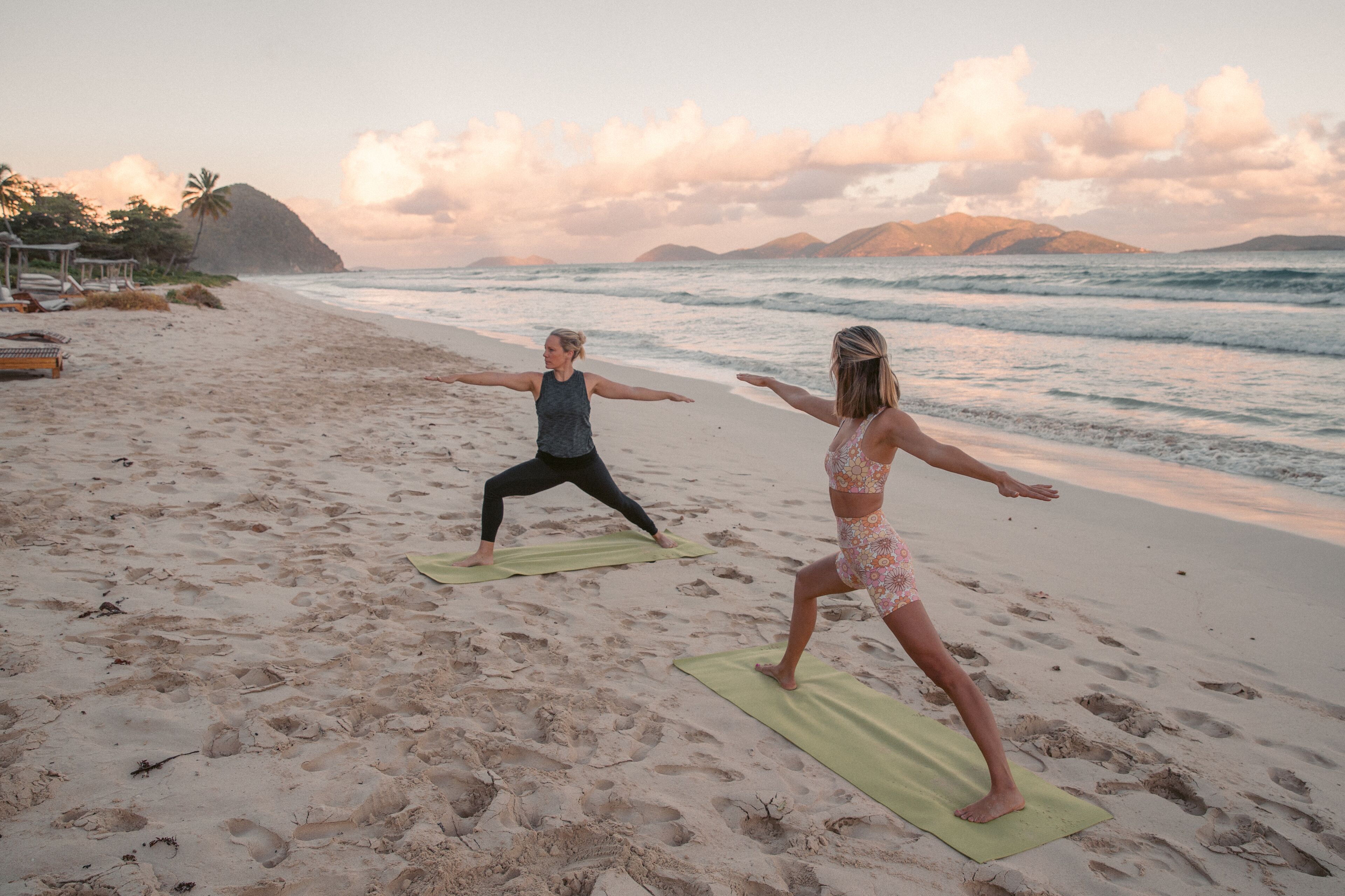 Dos personas practicando yoga sobre esterillas verdes en una playa arenosa con olas del océano al fondo.