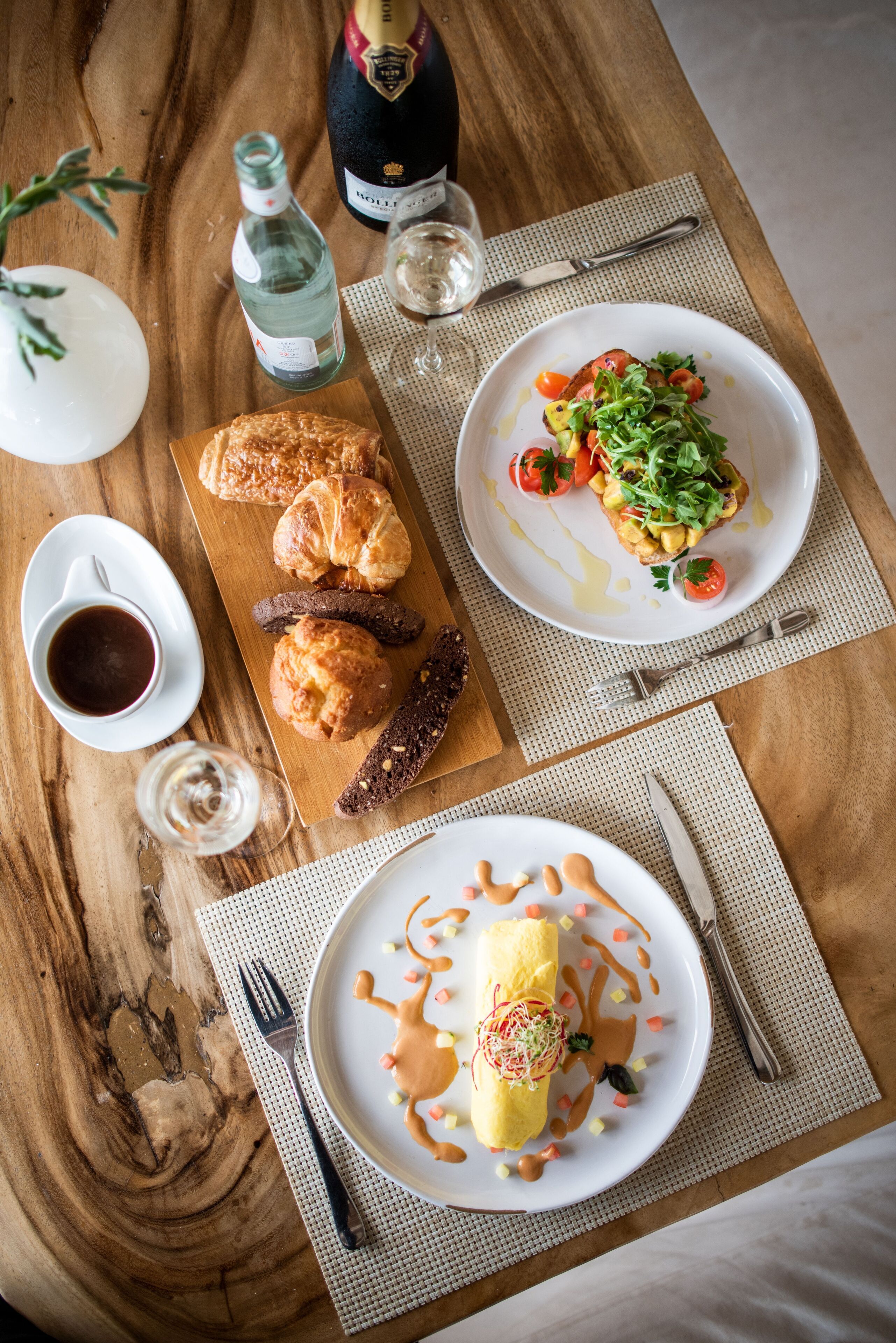 Et træbord med morgenmadsretter, blandt andet croissanter, en tallerken med æg og en champagneflaske.