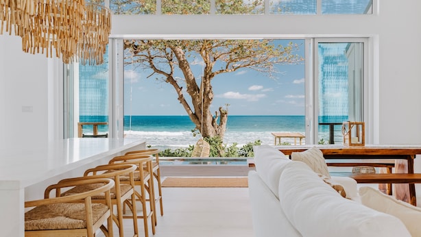 A modern interior with a large window overlooking the ocean, a wooden dining table, and a white sofa. - Long Bay Beach Resort (West End)