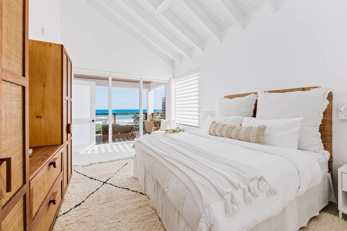 A bedroom with a large bed, a wooden wardrobe, and a view of the ocean through a sliding glass door.