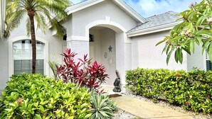 A residential building with a white exterior, a covered entrance, and a well-maintained garden with various plants and a palm tree. - Gorgeous Naples home with Saltwater pool in a gated community  (Naples)
