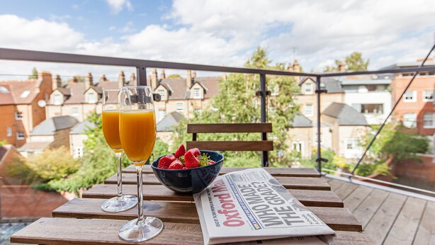Una mesa de madera con dos vasos de jugo de naranja, un cuenco de fresas y un periódico en un balcón con vistas a casas.