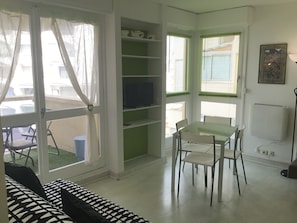 A room with a glass door leading to a balcony, a green bookshelf, a dining table with chairs, and a radiator on the wall.