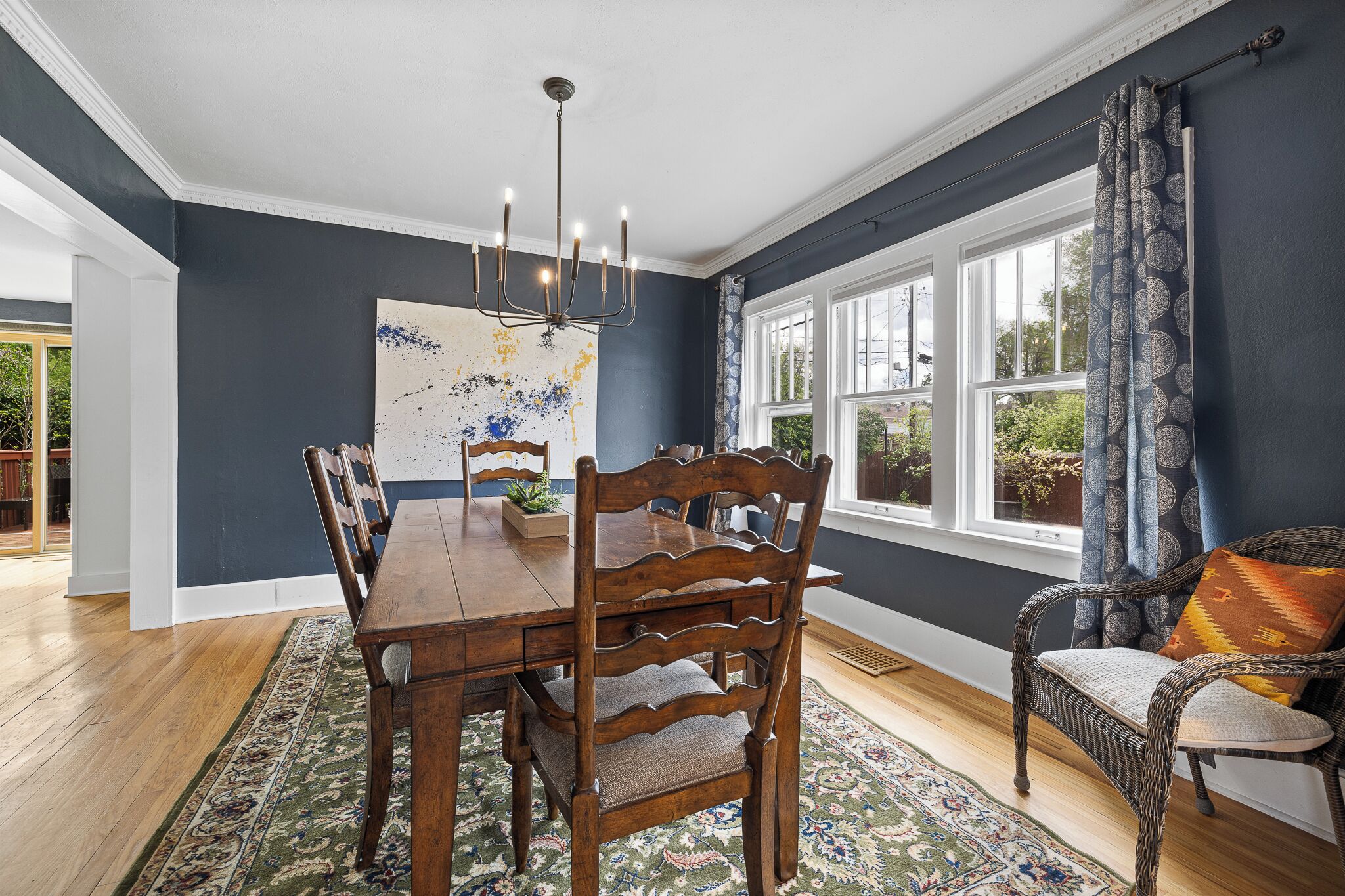A dining room with a wooden table and chairs, a chandelier, and a large window with curtains.