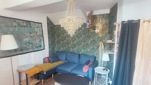 A living room with a blue sofa, a small round table, a floor lamp, and a decorative sunburst wall piece. - Cosy T2 apartment with river view, sleeps 2 (Rennes-les-Bains)