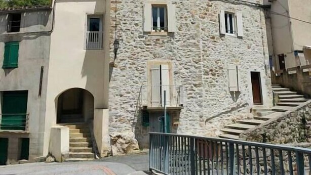 A stone building with a staircase leading to a second floor, a small door, and a metal railing in the foreground. - Cosy T2 apartment with river view, sleeps 2 (Rennes-les-Bains)