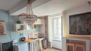 A compact kitchen with a chandelier, a refrigerator, a sink, and a window with curtains. - Cosy T2 apartment with river view, sleeps 2 (Rennes-les-Bains)