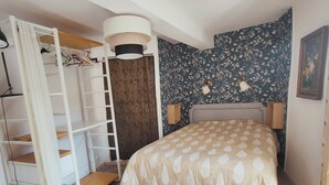 A bedroom with a bed, a wardrobe, a ceiling lamp, and floral wallpaper. - Cosy T2 apartment with river view, sleeps 2 (Rennes-les-Bains)