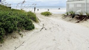 Ein sandiger Pfad am Strand, der zu einem weißen Gebäude mit horizontalen Lamellen mit Blick auf das Meer führt.