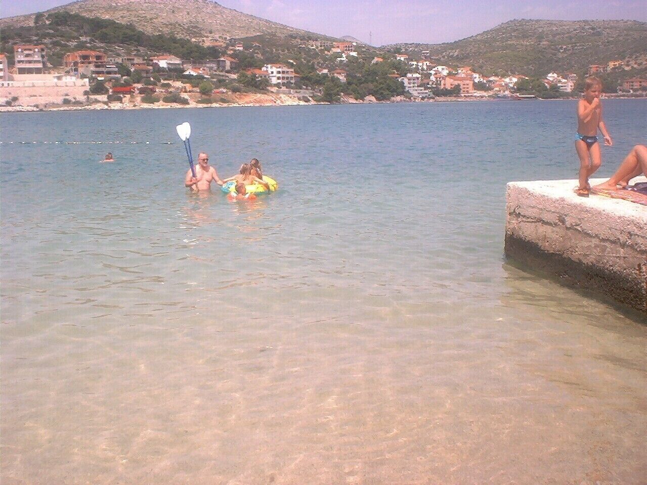 Eine Küstenansicht mit klarem Wasser, einem Steg und Gebäuden im Hintergrund.