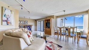 Uma sala de estar ampla com uma grande janela com vista para a praia, um sofá confortável e uma área de jantar com cadeiras.