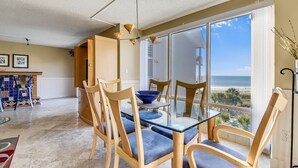 Uma sala de jantar com mesa de vidro e cadeiras de madeira, vista para a praia, e um teto com luminária diferenciada.