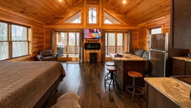 A log cabin interior with a large bed, a kitchen area, a dining table, and a TV in the center. - Malbec Cabin - 6 Guests - Hocking Hills - Hot Tub-Firepit-Wifi (Logan)