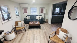 A living room with a dark leather sofa, two wicker chairs, a coffee table, and a kitchen visible through an open doorway. - Create Memories at our Modern Waterfront 1 BR Apartment: Hike, Sip Wine, Relax!  (Wallkill)
