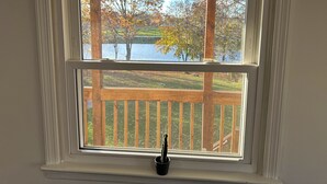 A window with a view of a lake and trees, a wooden deck, and a potted plant on the windowsill. - Create Memories at our Modern Waterfront 1 BR Apartment: Hike, Sip Wine, Relax!  (Wallkill)