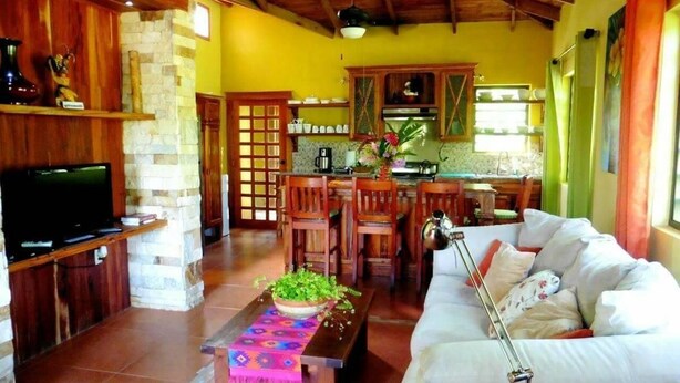 A cozy living room with a sofa, a coffee table, a television, and a kitchen area in the background. - Aliana's Villas - Private Farm Home on the River (Gold Standard Certified) (San Ignacio)
