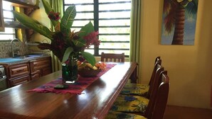 A wooden dining table with a floral cushion, a vase with flowers, and a bowl of fruit. - Aliana's Villas - Private Farm Home on the River (Gold Standard Certified) (San Ignacio)