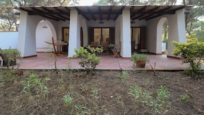 Uma área externa coberta com teto de madeira, paredes brancas e chão de azulejos. Várias plantas em vasos e uma rede.