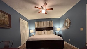 Quarto com cama, duas luminárias de cabeceira, ventilador de teto, porta e uma cadeira.