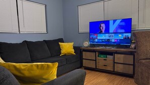 Uma sala de estar com televisão exibindo um programa, um sofá com almofadas amarelas e um armário de madeira com gavetas.