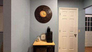 Quarto com porta branca, relógio na parede, cafeteira e pequena mesa com micro-ondas.