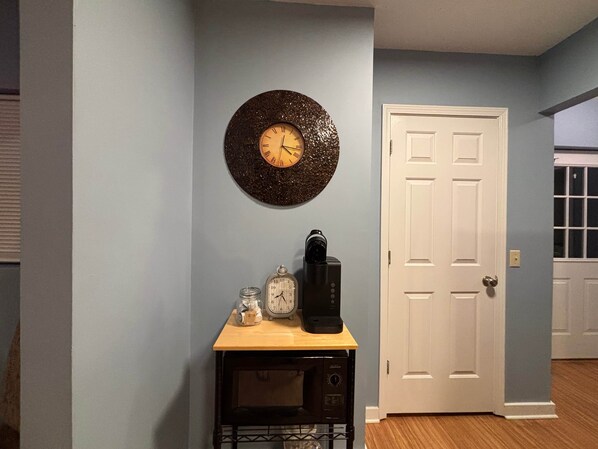A room with a white door, a wall clock, a coffee maker, and a small table with a microwave. - Woodward Villa D (Dahlonega)
