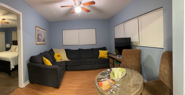 A living room with a dark sofa, a round coffee table with magazines, a ceiling fan, and a bed in the background.