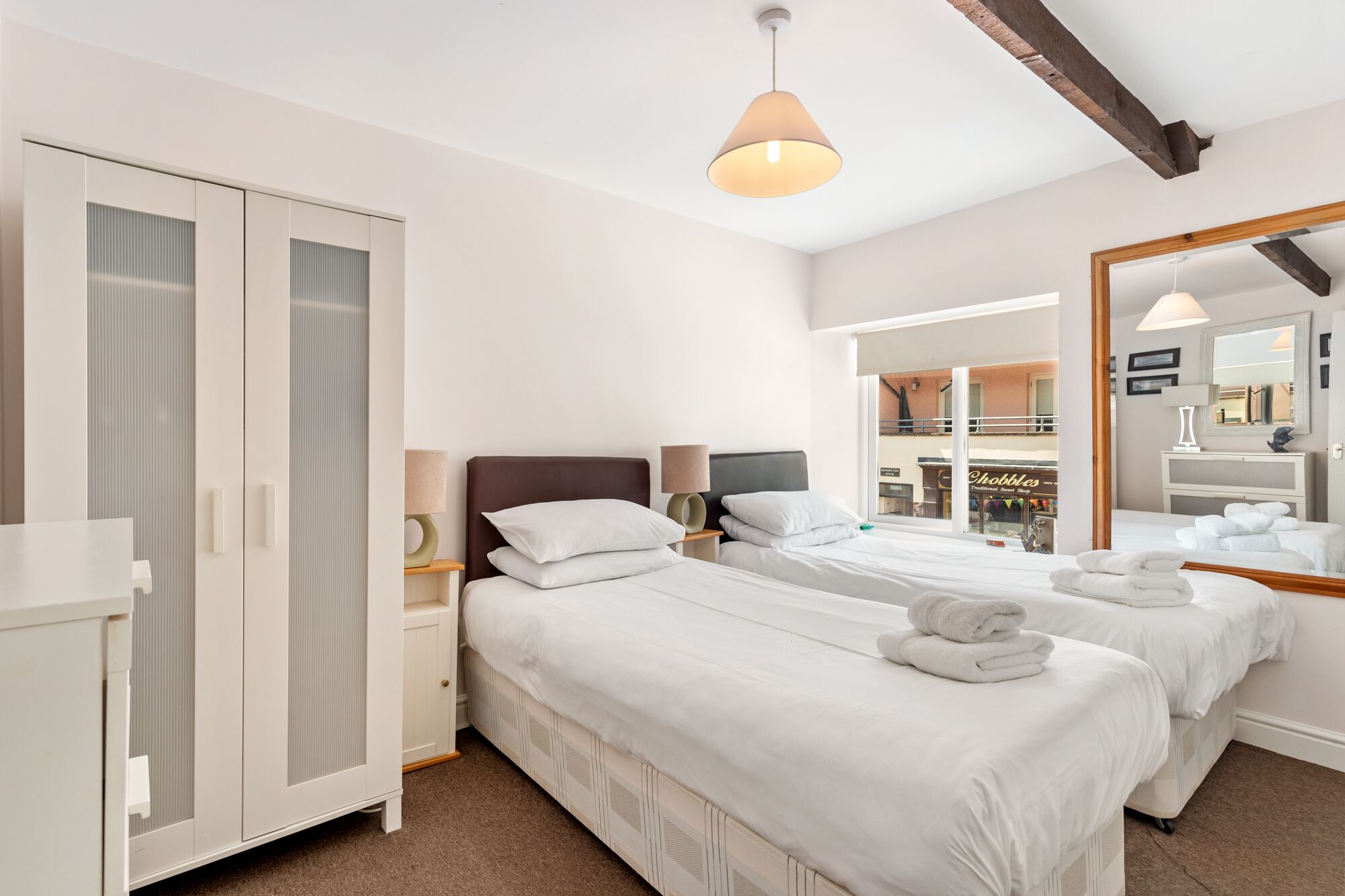 A hotel room with two beds, a wardrobe, a mirror, and a view of a traditional sweet shop.