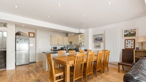 A modern kitchen with wooden cabinets, a built-in oven, and a dining area with a wooden table and chairs. - Kilmore - Direct Beach Access, Sea Views (Saundersfoot)