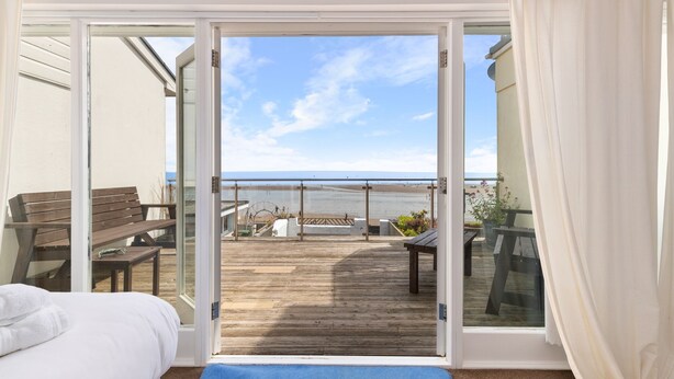 A room with a bed, a wooden bench, and a view of a beach through a glass door. - Kilmore - Direct Beach Access, Sea Views (Saundersfoot)