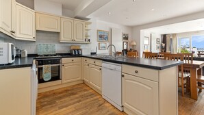 A modern kitchen with white cabinetry, a dishwasher, and a wooden dining area with a view. - Kilmore - Direct Beach Access, Sea Views (Saundersfoot)