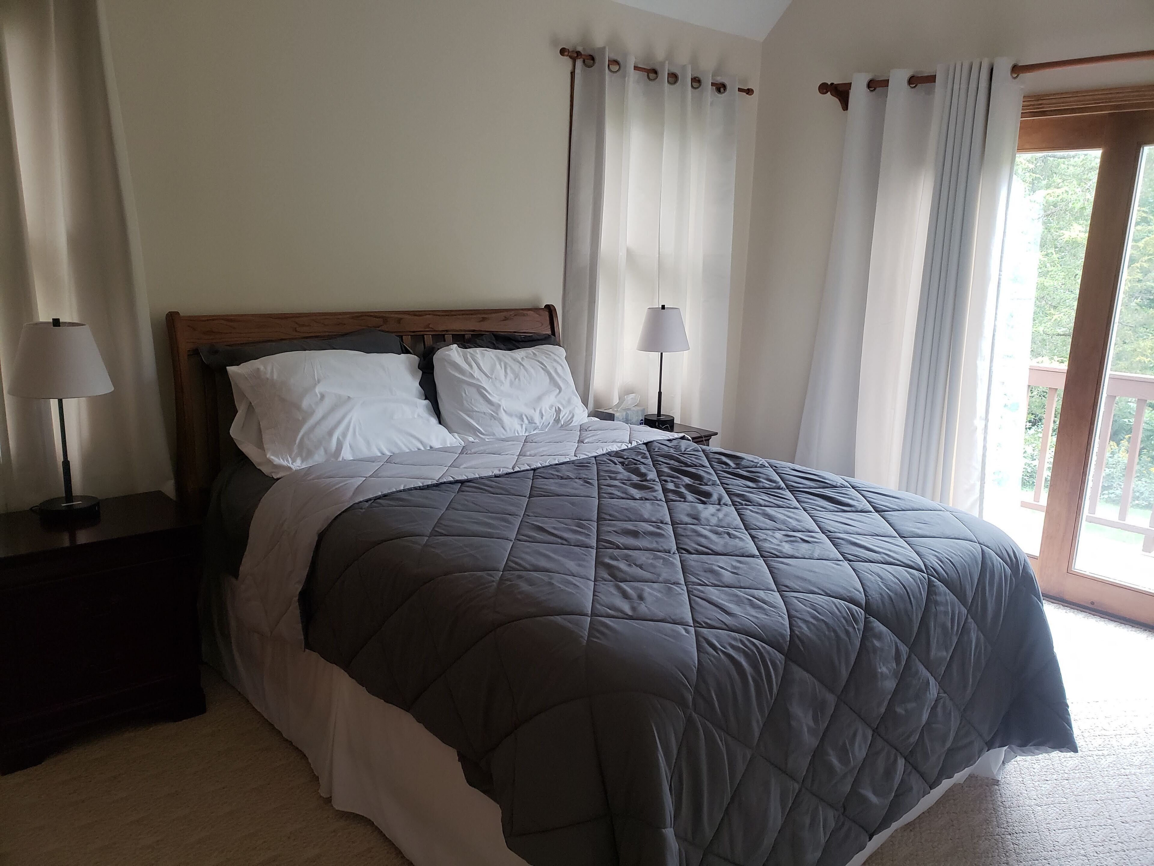 A neatly made bed with a dark quilt, two bedside lamps, and a window with curtains.