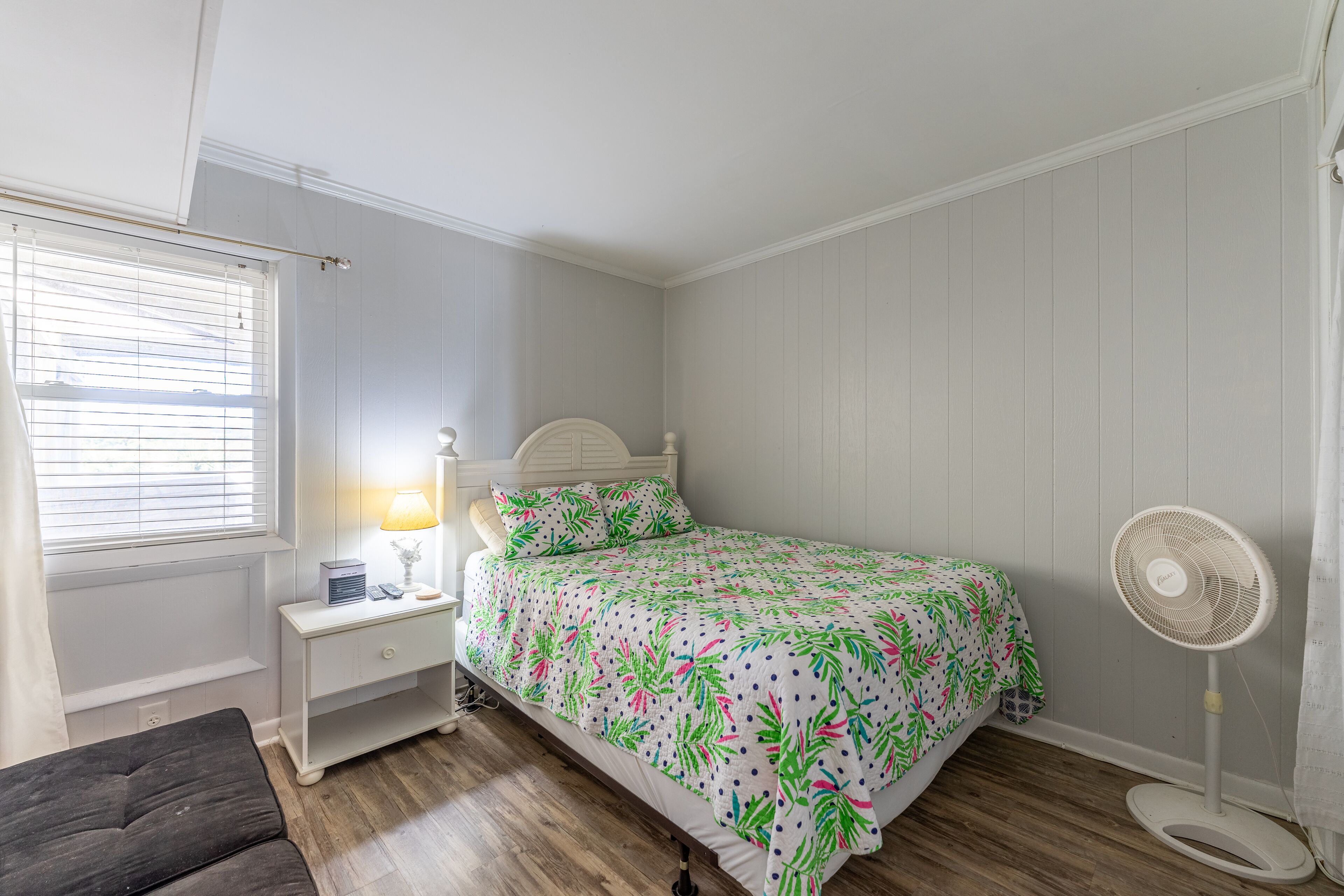 Un dormitorio con cama, mesita de noche, lámpara y ventilador de pie.